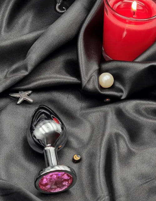 Side view of the polished steel butt plug beside a candle, showing smooth tapered tip and flared jewel base.