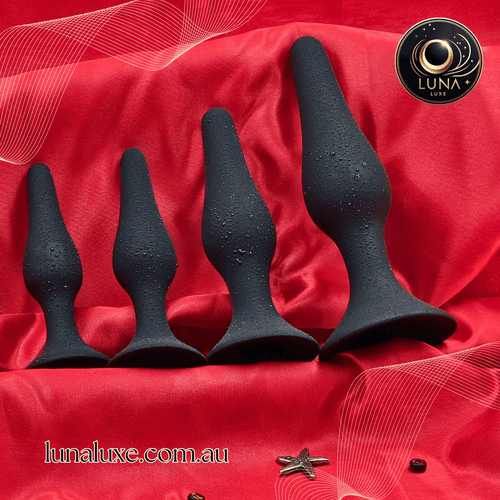 Black silicone anal plug set on red satin, four sizes with tapered tips and flared safety bases.