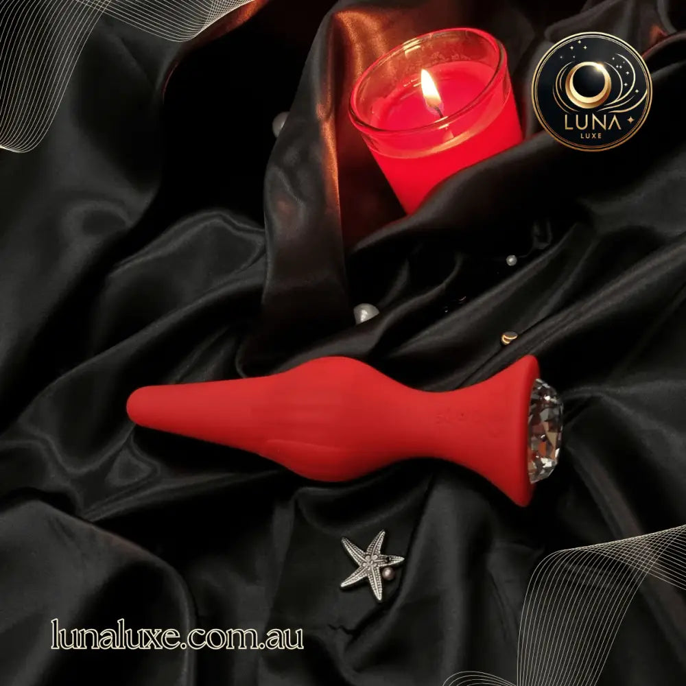 Red silicone jeweled anal plug on black satin, tapered bulb with faceted crystal base.