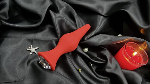 Angled view of the red butt plug on satin, showing smooth silicone and flared gem stopper.